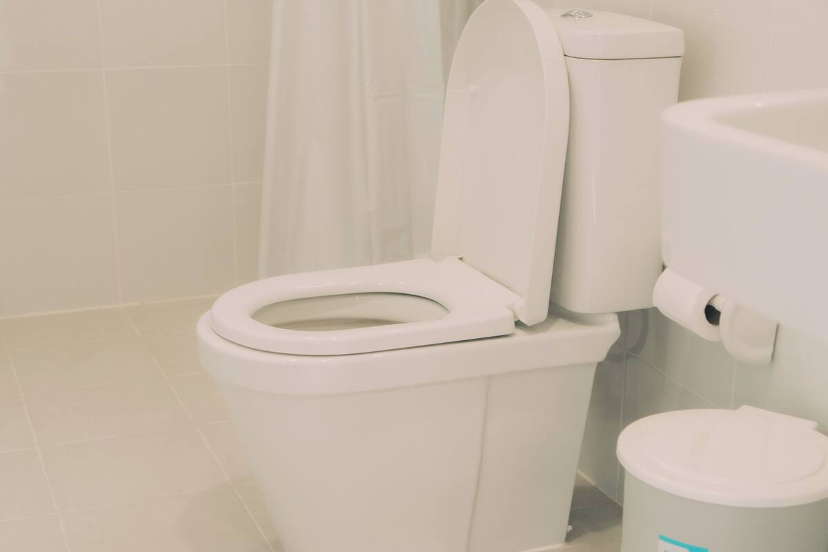 A toilet with a closed lid, a white trash bin, and a shower curtain in a bathroom with white tiled walls and floor.