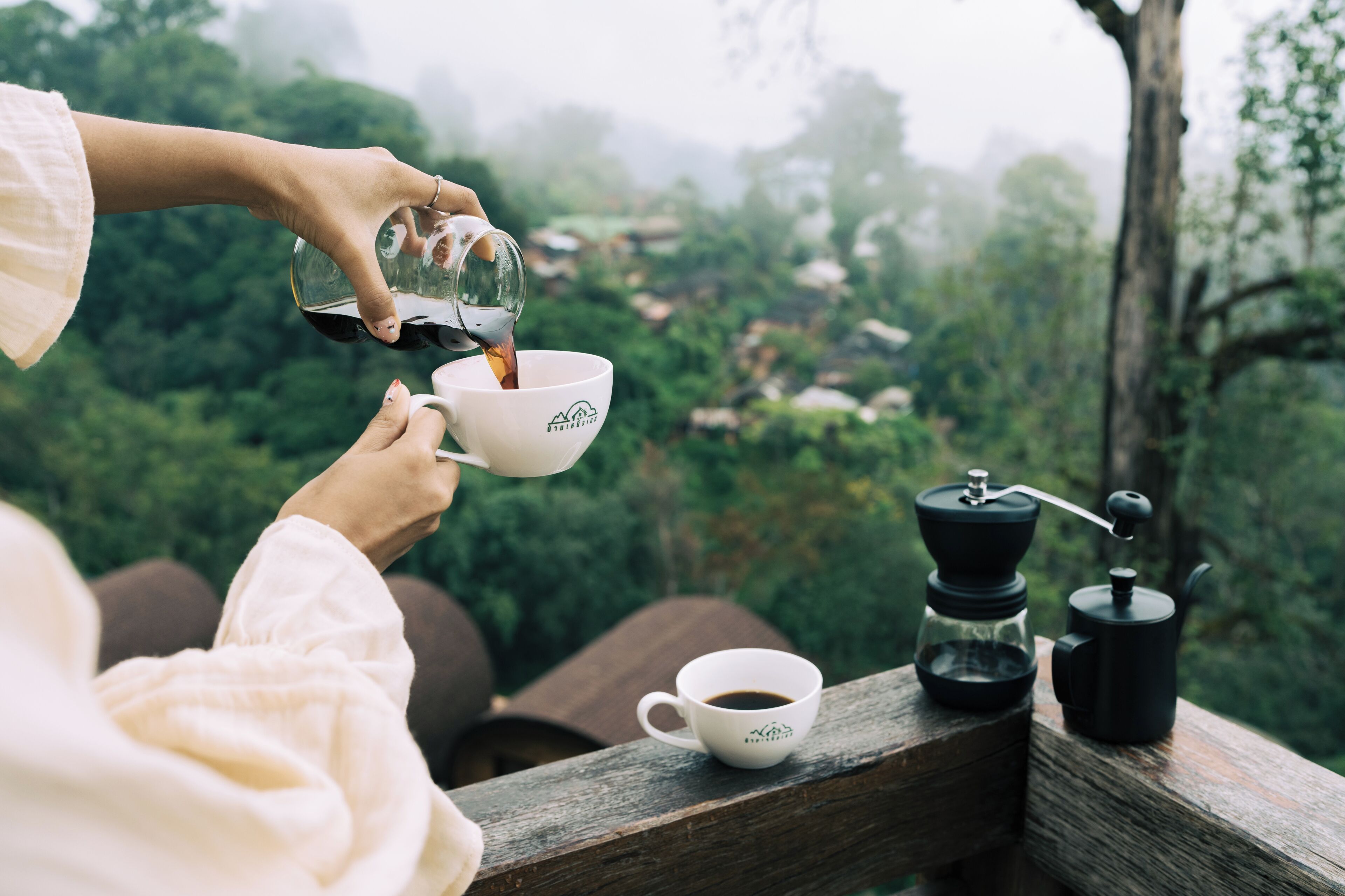 A person pouring coffee into a cup, a coffee grinder, and a cup on a wooden railing with a view of greenery.