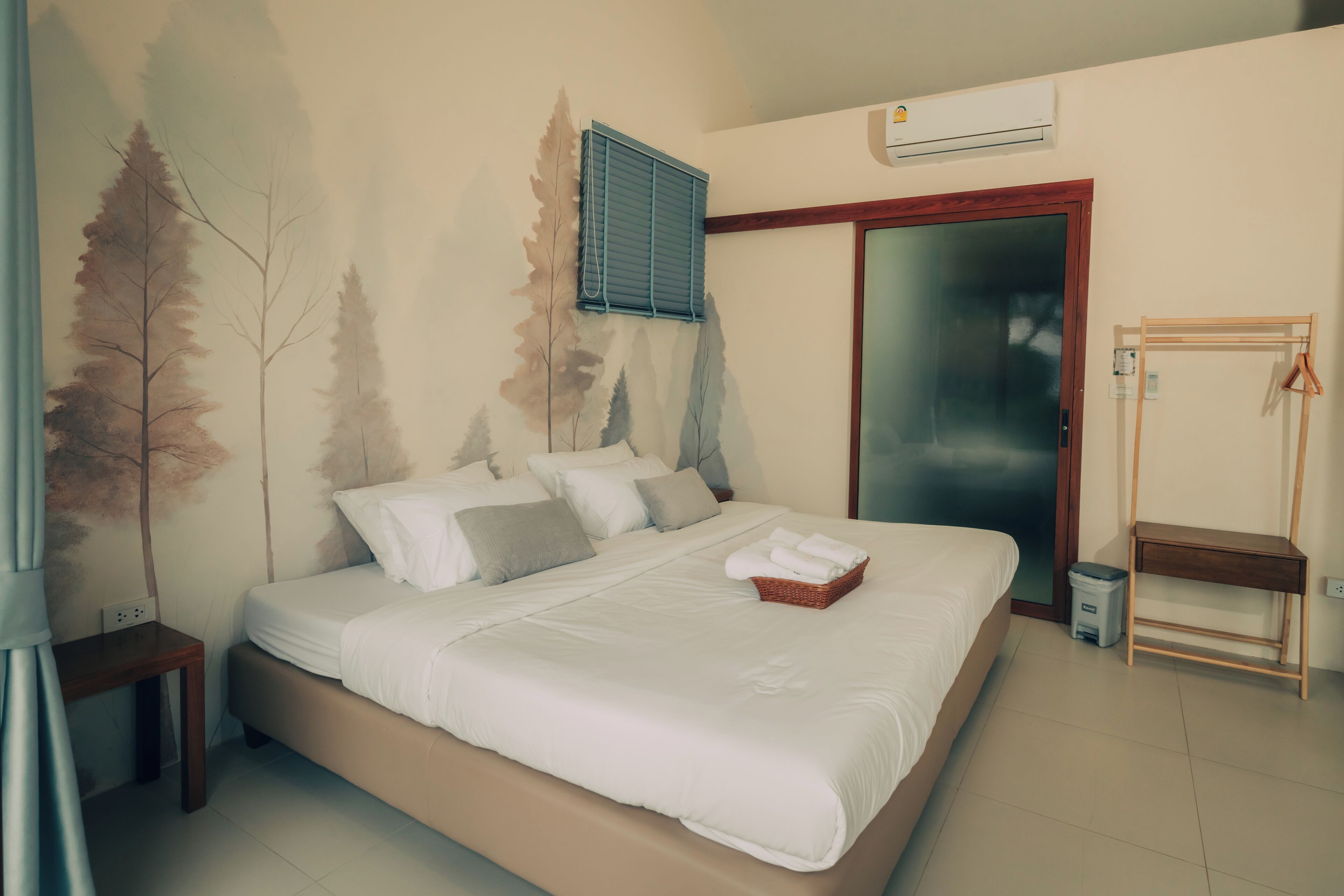 A bedroom with a bed, a wall-mounted air conditioner, a wooden chair, and a small trash bin.