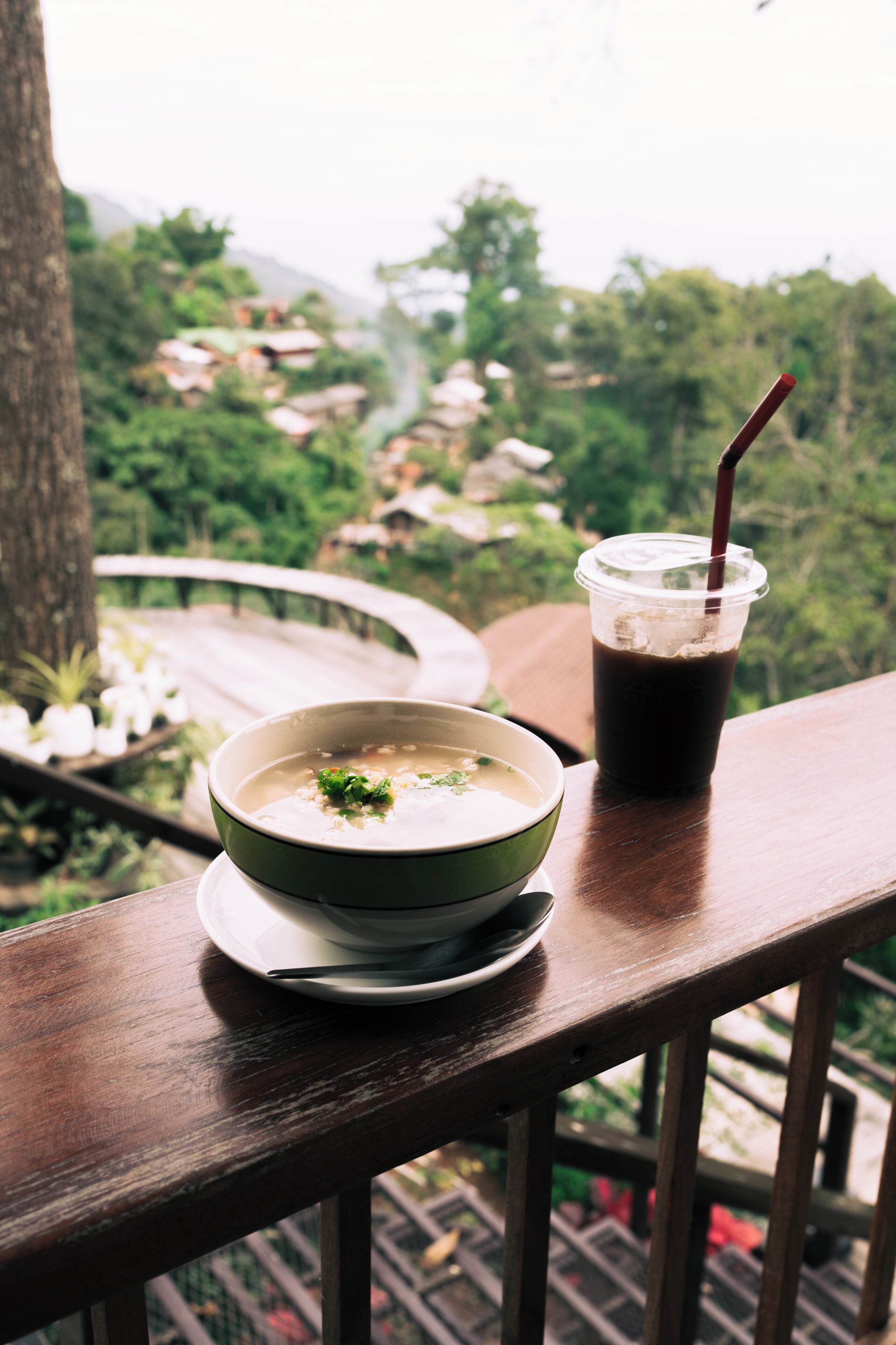 Una mesa con un tazón de sopa y una bebida en una terraza de madera con vistas a un paisaje exuberante.