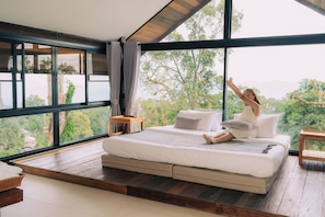 A bedroom with a large window, a bed, a nightstand, and a view of trees.