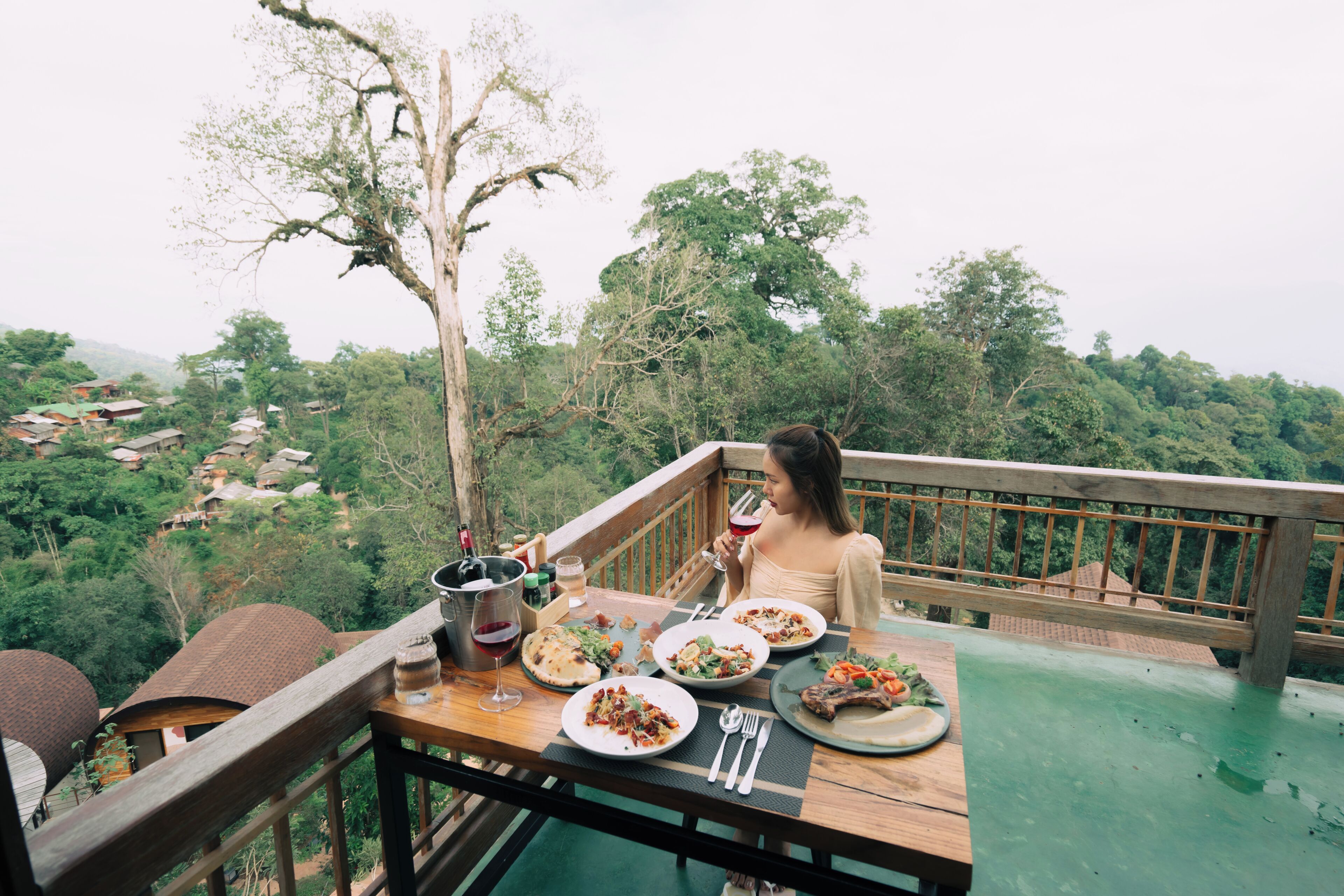 A dining table with plates of food, a wine glass, and cutlery on a balcony overlooking a forested area.