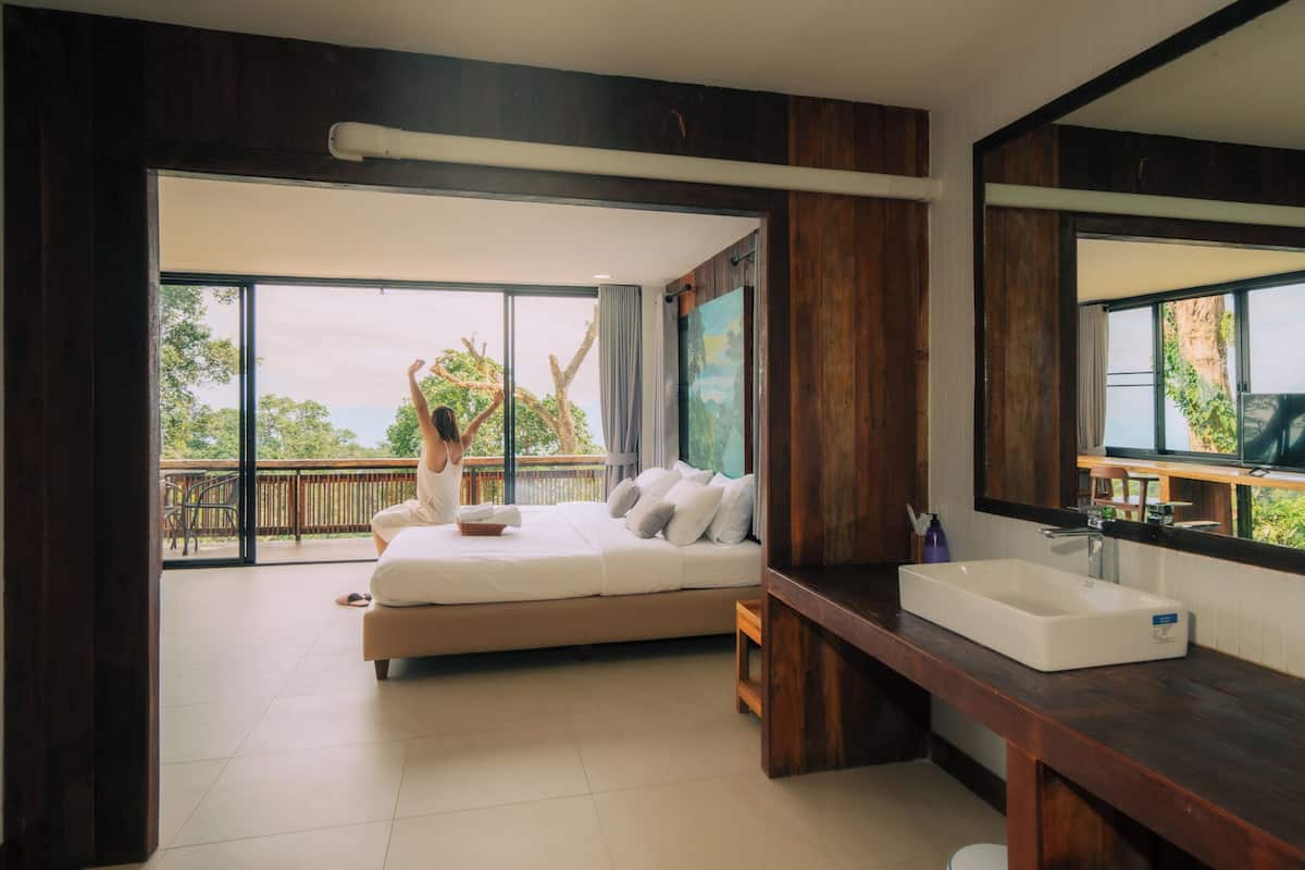A bedroom with a large bed, a wooden vanity, and a balcony with a view of trees.