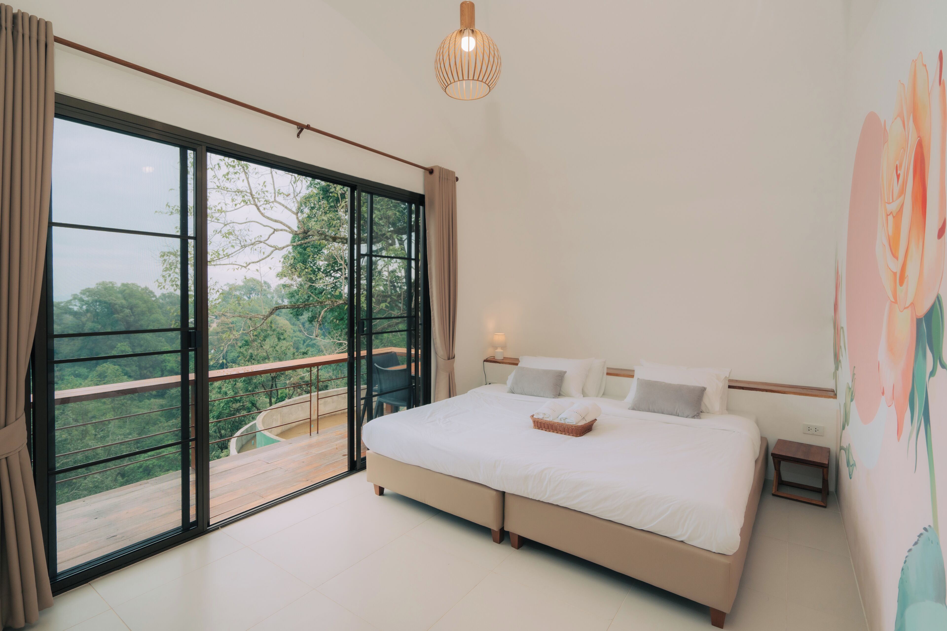 A bedroom with a large bed, a nightstand, a wall-mounted lamp, a window with curtains, and a balcony with a view of trees.