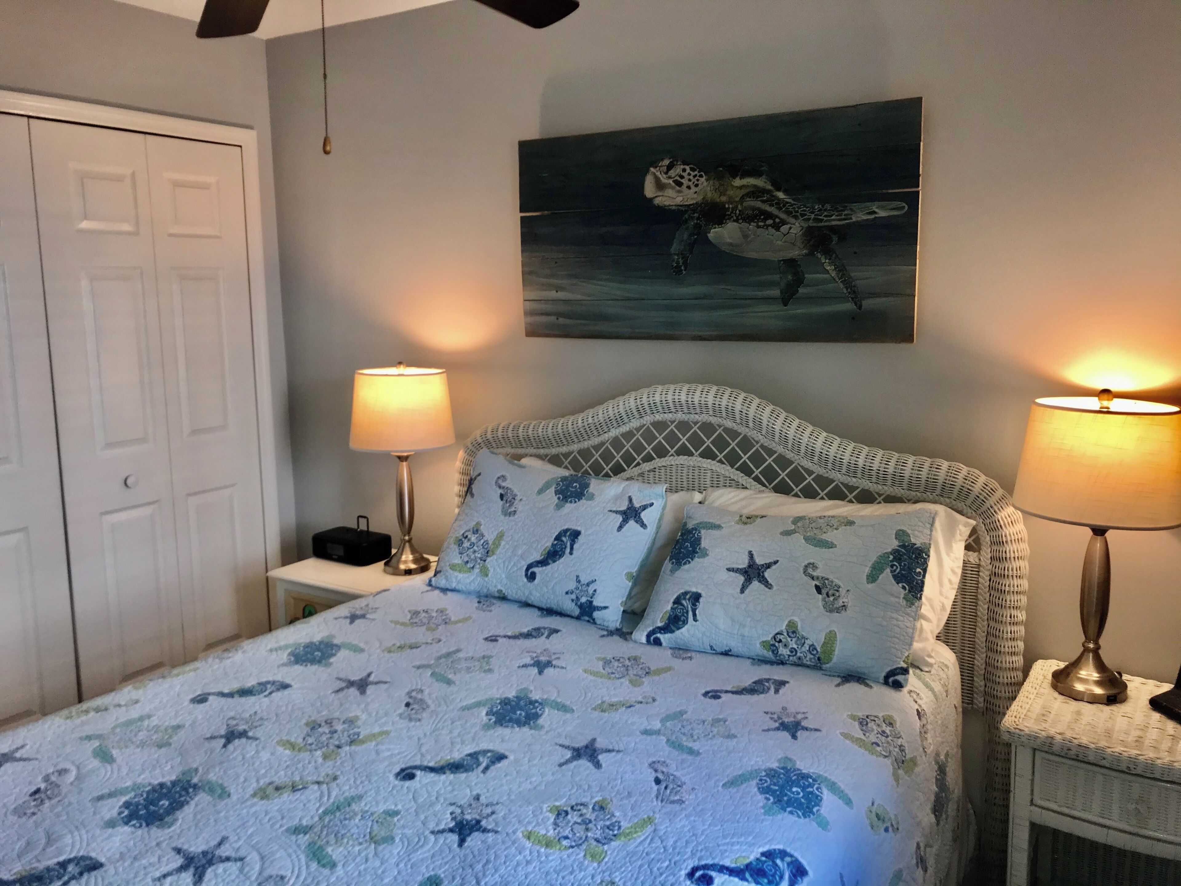A bedroom with a bed featuring a sea-themed duvet, two bedside lamps, a white door, and a wall-mounted painting of a sea turtle.