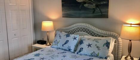 A bedroom with a bed featuring a sea-themed duvet, two bedside lamps, a white door, and a wall-mounted painting of a sea turtle.