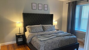 Quarto com cabeceira escura, roupa de cama estampada, duas luminárias de cabeceira e uma janela com cortinas.