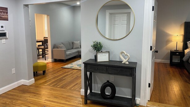 Uma sala de estar moderna com um sofá cinzento, um espelho redondo, uma pequena mesa de apoio com uma planta e uma decoração em formato de coração, e piso de madeira.