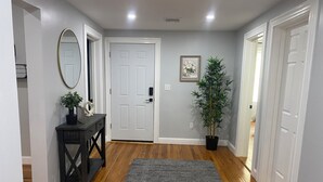Um corredor com piso de madeira, um tapete cinza, um console de madeira escura com uma planta, um espelho e uma porta com moldura de imagem.