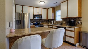 Cocina compacta con refrigerador, microondas, estufa y cafetera. Hay dos taburetes de bar y una ventana con persianas.