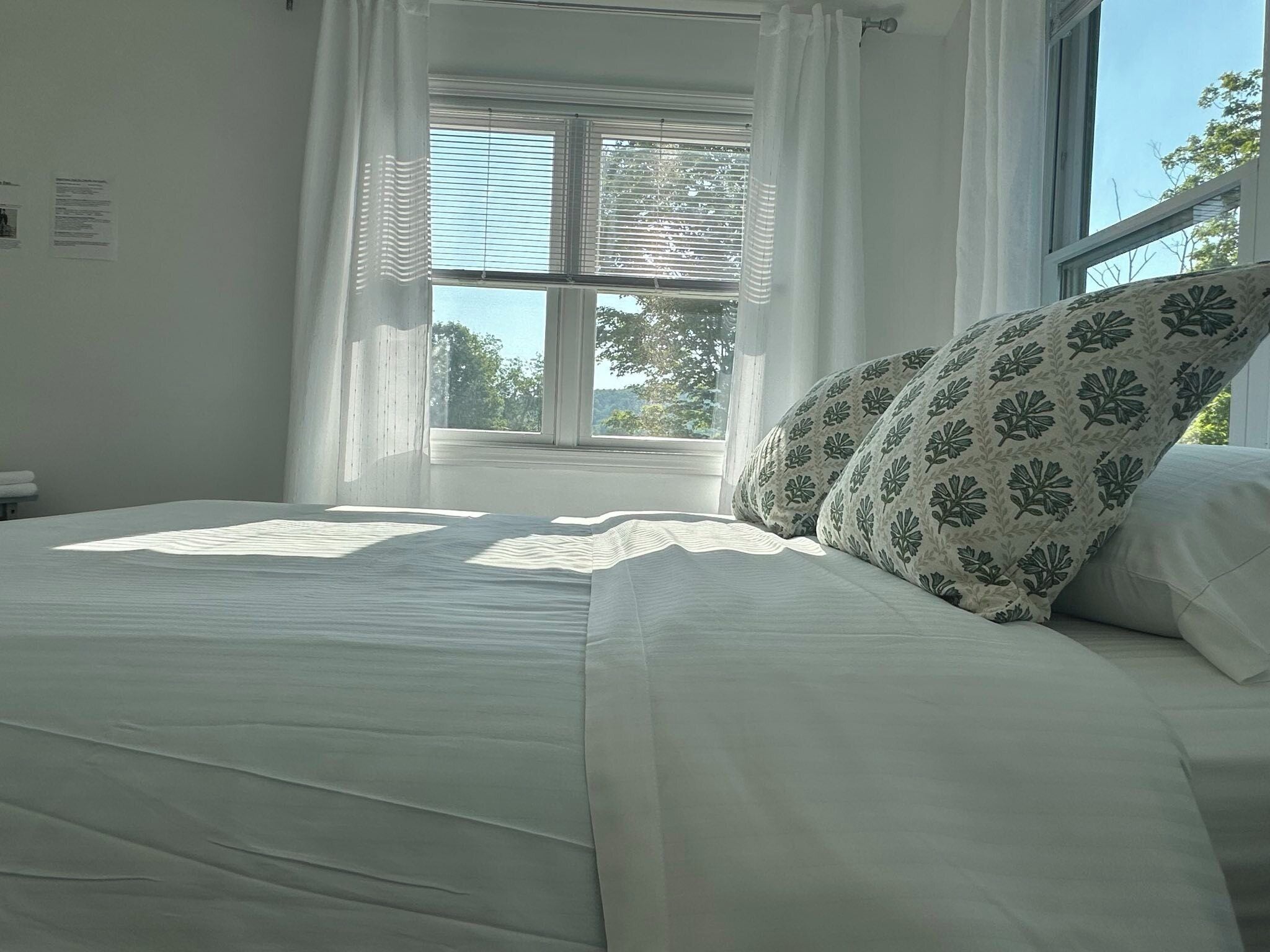 A bedroom with a bed, white curtains, and a window with blinds.