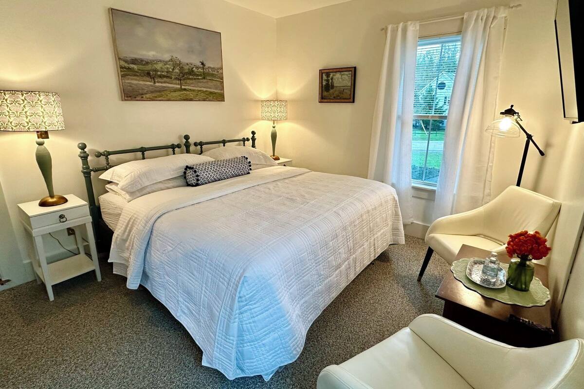 A bedroom with a bed, bedside lamps, a chair, a window with curtains, and artwork on the walls.