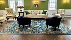 Ein Wohnzimmer mit grüner Wand, zwei Fenstern, einem beigen Sofa, einem floralen Teppich und einem schwarzen Sessel.