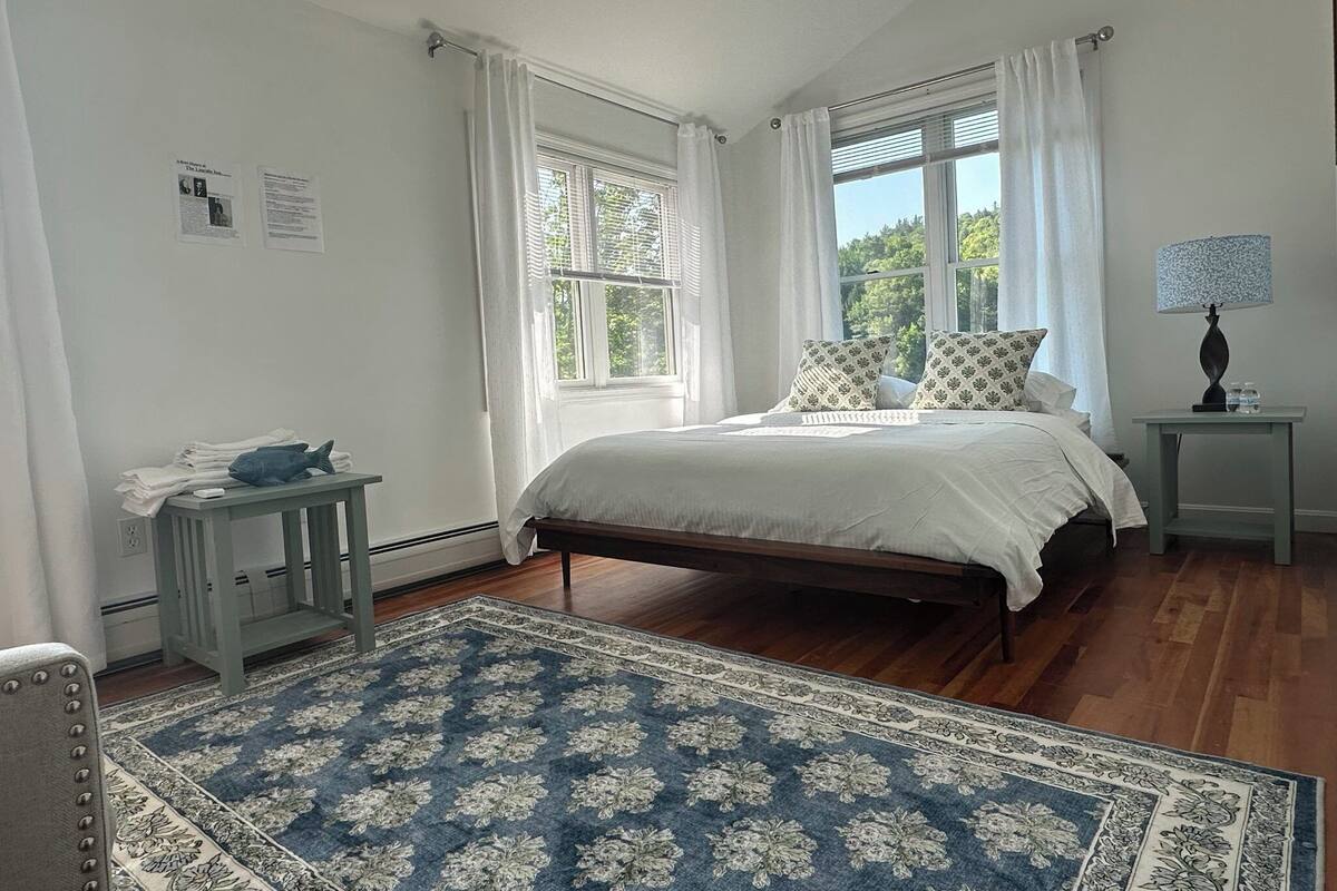 A bedroom with a bed, a nightstand, a lamp, a window with curtains, a blue patterned rug, and a small table with folded towels.
