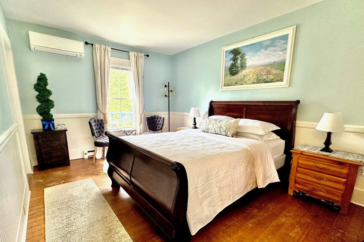 A bedroom with a large bed, a nightstand, a lamp, a chair, a small table, and a window with curtains.