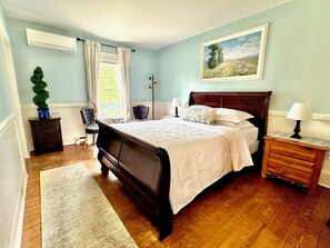 A bedroom with a large bed, a nightstand, a lamp, a chair, a small table, and a window with curtains. - Lincoln Inn & Gallery (Woodstock)