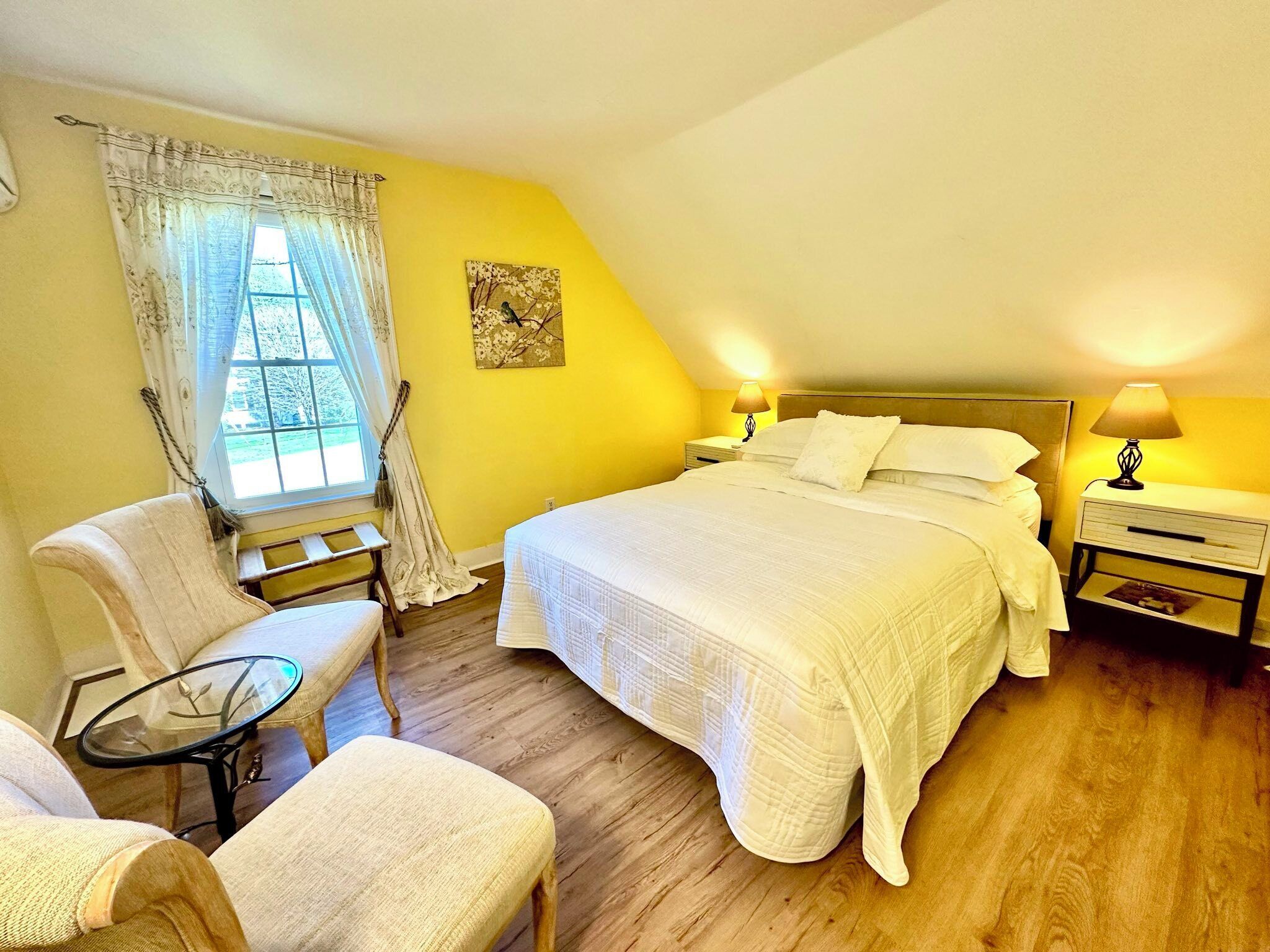 A bedroom with a bed, two bedside tables with lamps, a chair, a small table, and a window with curtains.