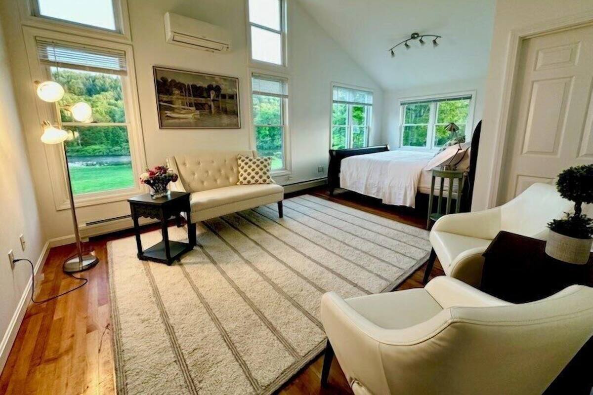 A spacious room with a large bed, a sofa, and a chair, featuring large windows and a view of greenery outside.