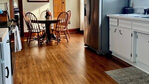 Una cocina con suelo de madera, refrigerador de acero inoxidable, armarios blancos y un área de comedor con sillas y mesa de madera.