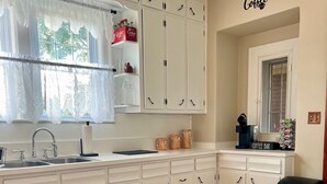 Una cocina con armarios blancos, una barra de café y un ventanal con cortinas de encaje.