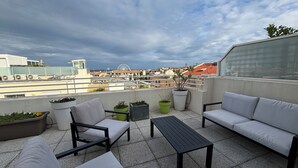 Terraza en la azotea con mobiliario exterior, plantas en macetas y vistas a la ciudad con una noria a lo lejos.