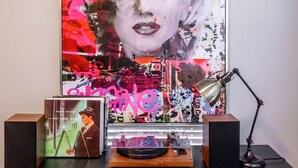 Un cartel muestra a una persona de cabello blanco y labios rojos, un escritorio con una lámpara y un altavoz.
