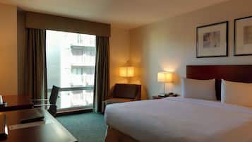 Une chambre d’hôtel avec un grand lit, un bureau avec une chaise, une fenêtre avec des rideaux, deux lampes de chevet et des tableaux encadrés au mur.