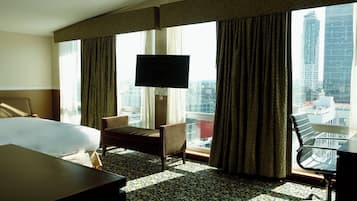 A hotel room with a large bed, a desk with a chair, a television, and a view of the cityscape through large windows.