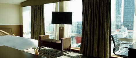 A hotel room with a large bed, a desk with a chair, a television, and a view of the cityscape through large windows.