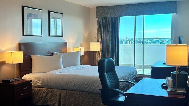 Quarto de hotel com uma cama, duas mesas de cabeceira com abajures, uma escrivaninha com cadeira e uma janela grande com vista para a cidade.