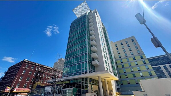 A modern building with a glass facade, a flat roof, and a white base. - LIC Manhattan View Hotel (Long Island City)
