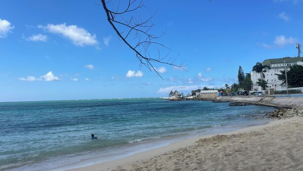 Una playa con agua azul cristalina, una orilla arenosa y edificios al fondo.