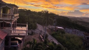 Un edificio de dos pisos con un balcón y vistas a un camino de tierra que se extiende hacia un paisaje montañoso.