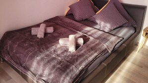Een keurig opgemaakt bed met een paars dekbed en kussens, een houten vloer en twee ingelijste foto's aan de muur.