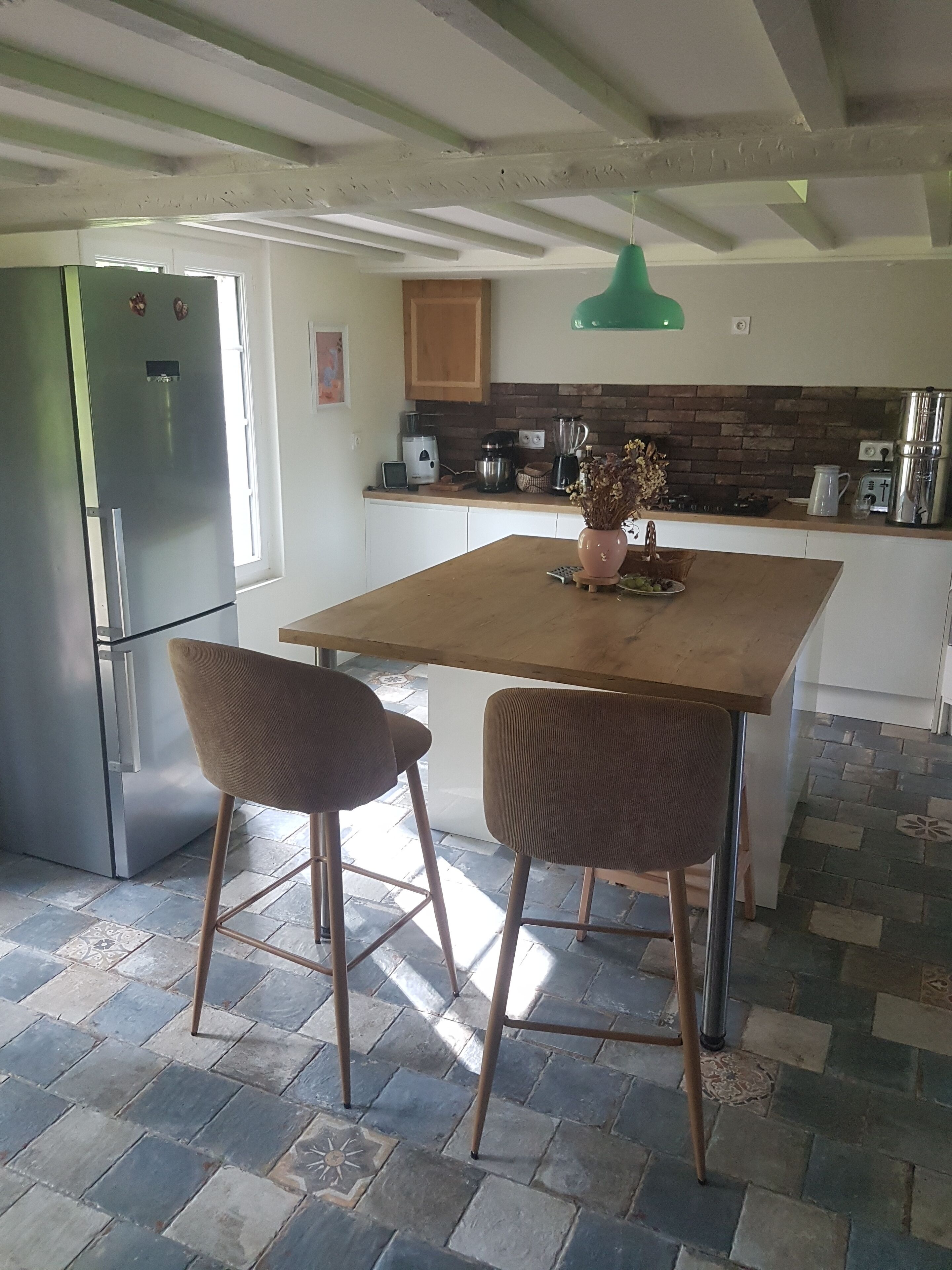 Une cuisine avec une table en bois, deux chaises, un réfrigérateur, une machine à café et un mur recouvert de briques.