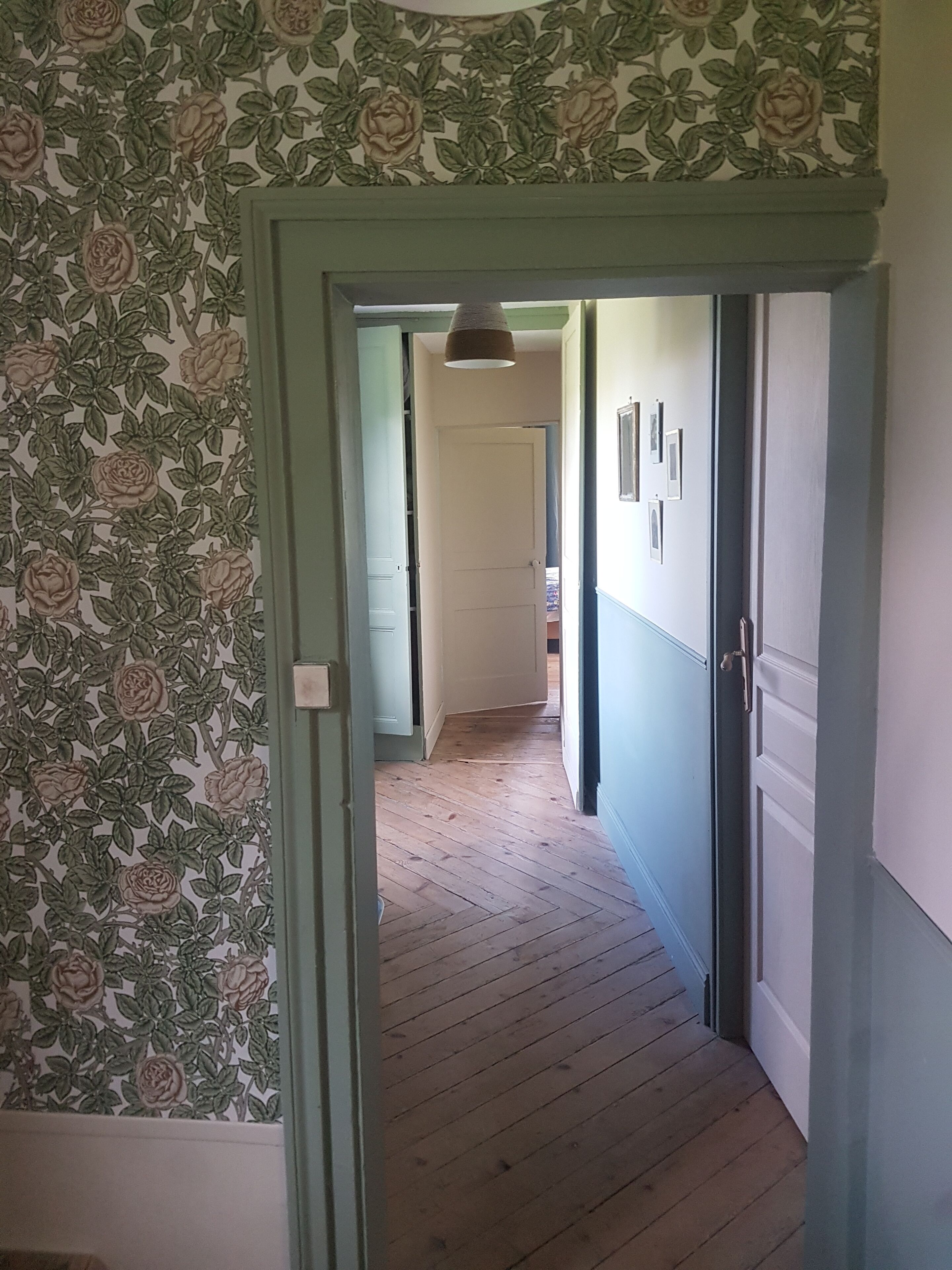 Un pasillo con papel tapiz floral, suelo de madera y una puerta que da a otra habitación.