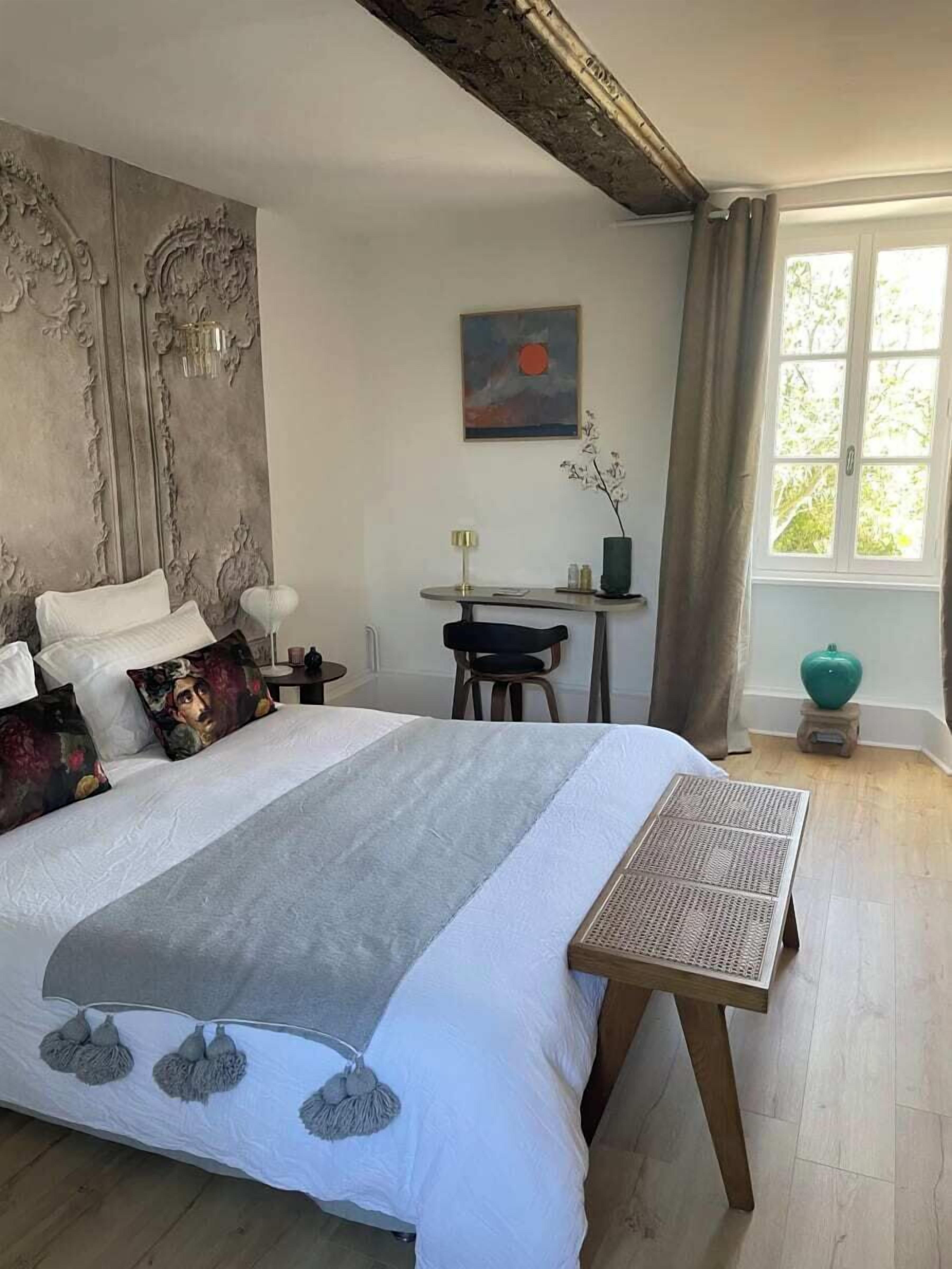 Et soveværelse med en stor seng, et lille bord, en stol, et billede og et vindue med gardiner.