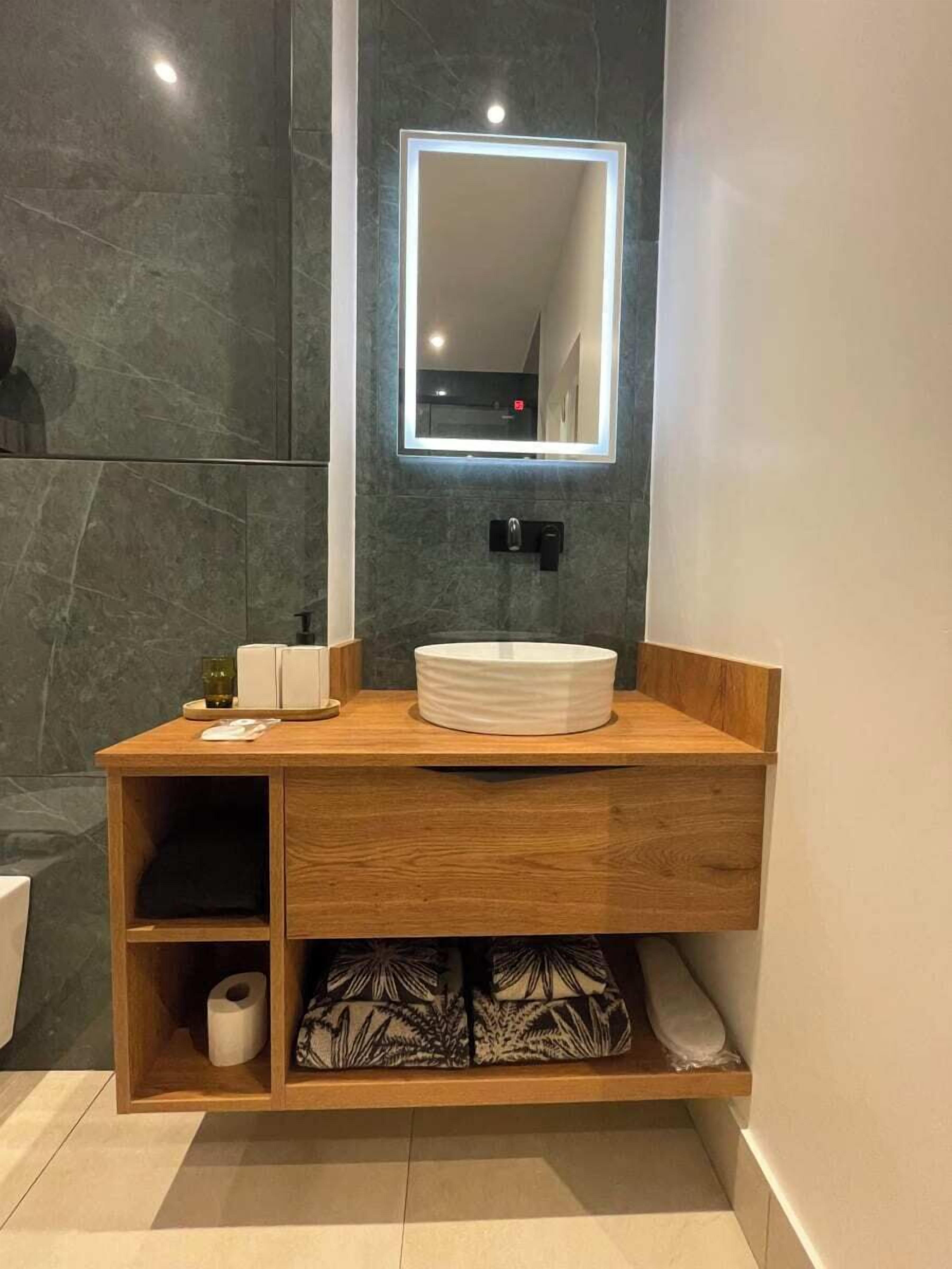 A modern bathroom with a wooden vanity, a round sink, and a mirror with built-in lighting.