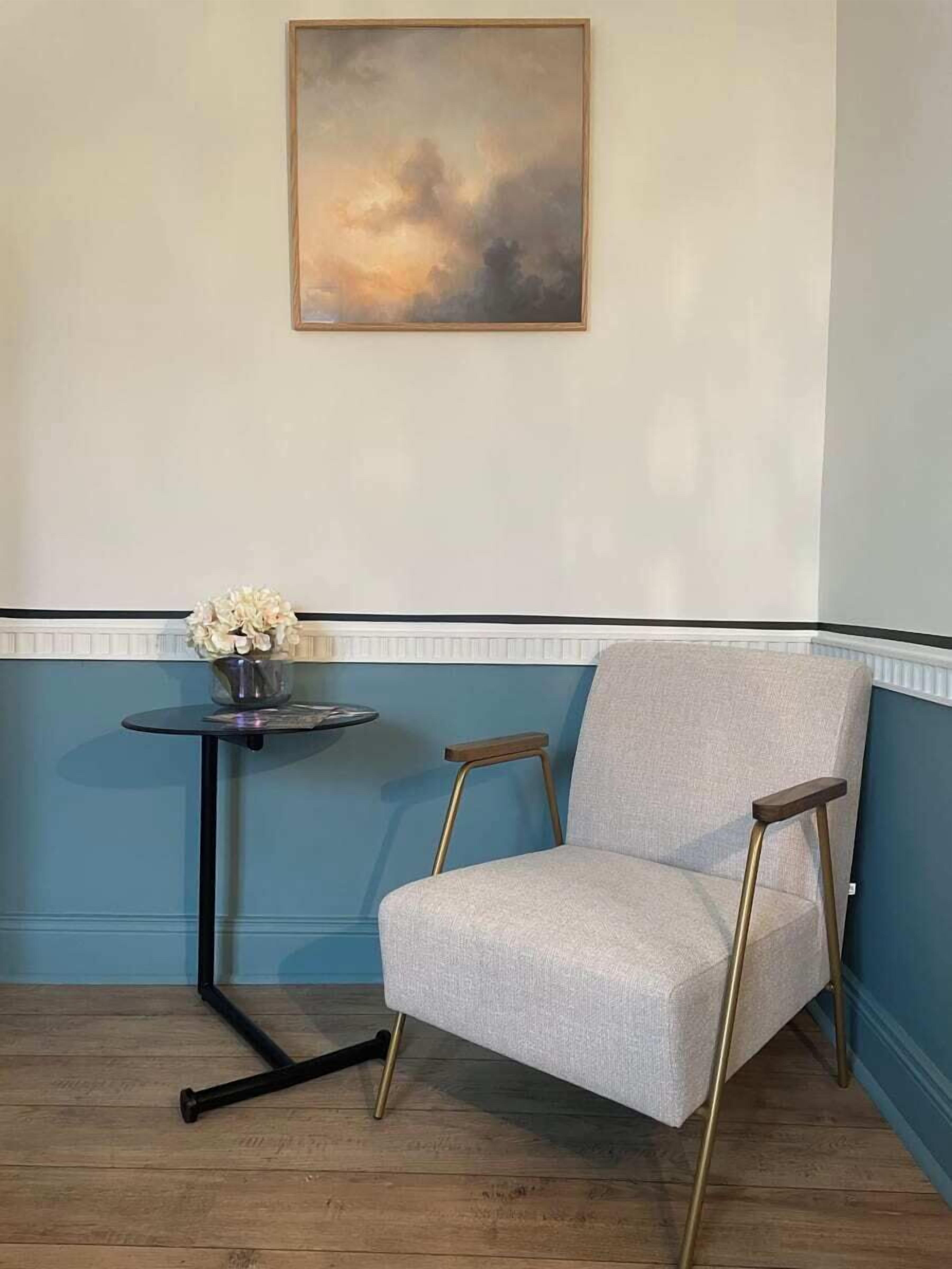 Quarto de canto com lambris azul, uma poltrona cinza com pés dourados, uma mesa redonda pequena com um vaso de flores e um quadro na parede.