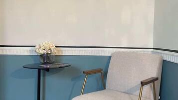 Quarto de canto com lambris azul, uma poltrona cinza com pés dourados, uma mesa redonda pequena com um vaso de flores e um quadro na parede.