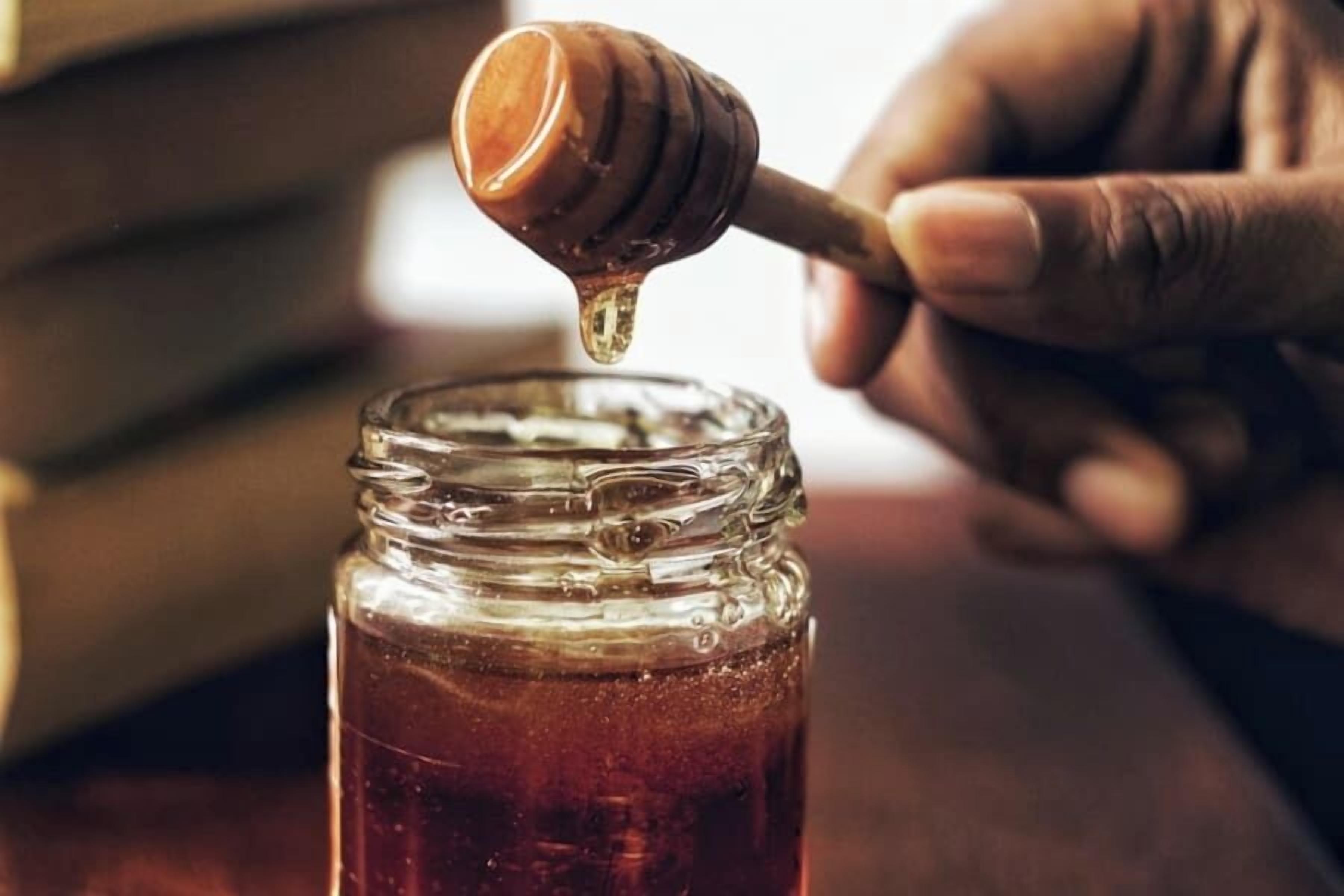 A hand holding a honey dipper dripping honey into a jar.