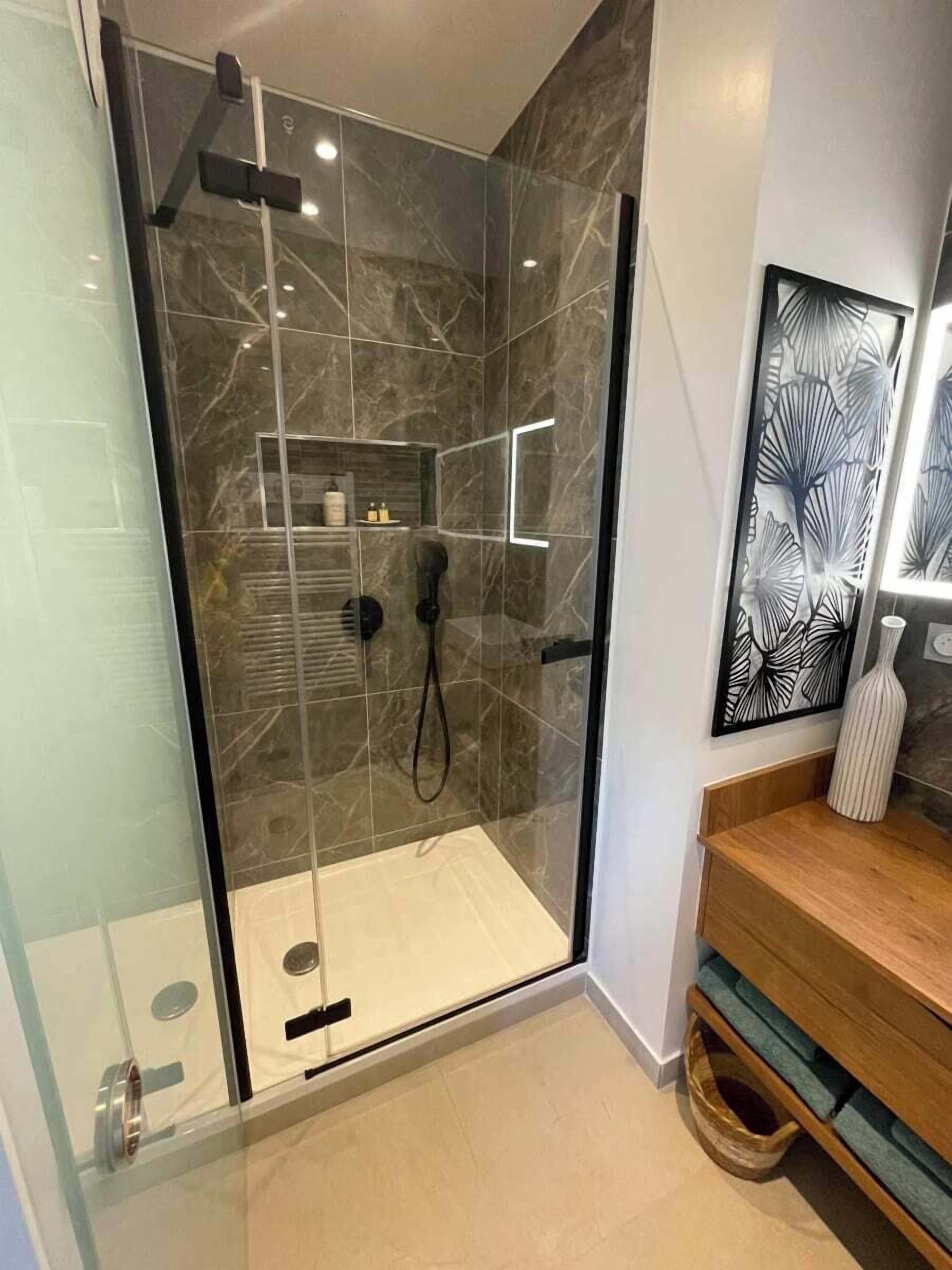 A modern bathroom with a glass-enclosed shower, marble tiles, a wooden vanity, and a decorative mirror.