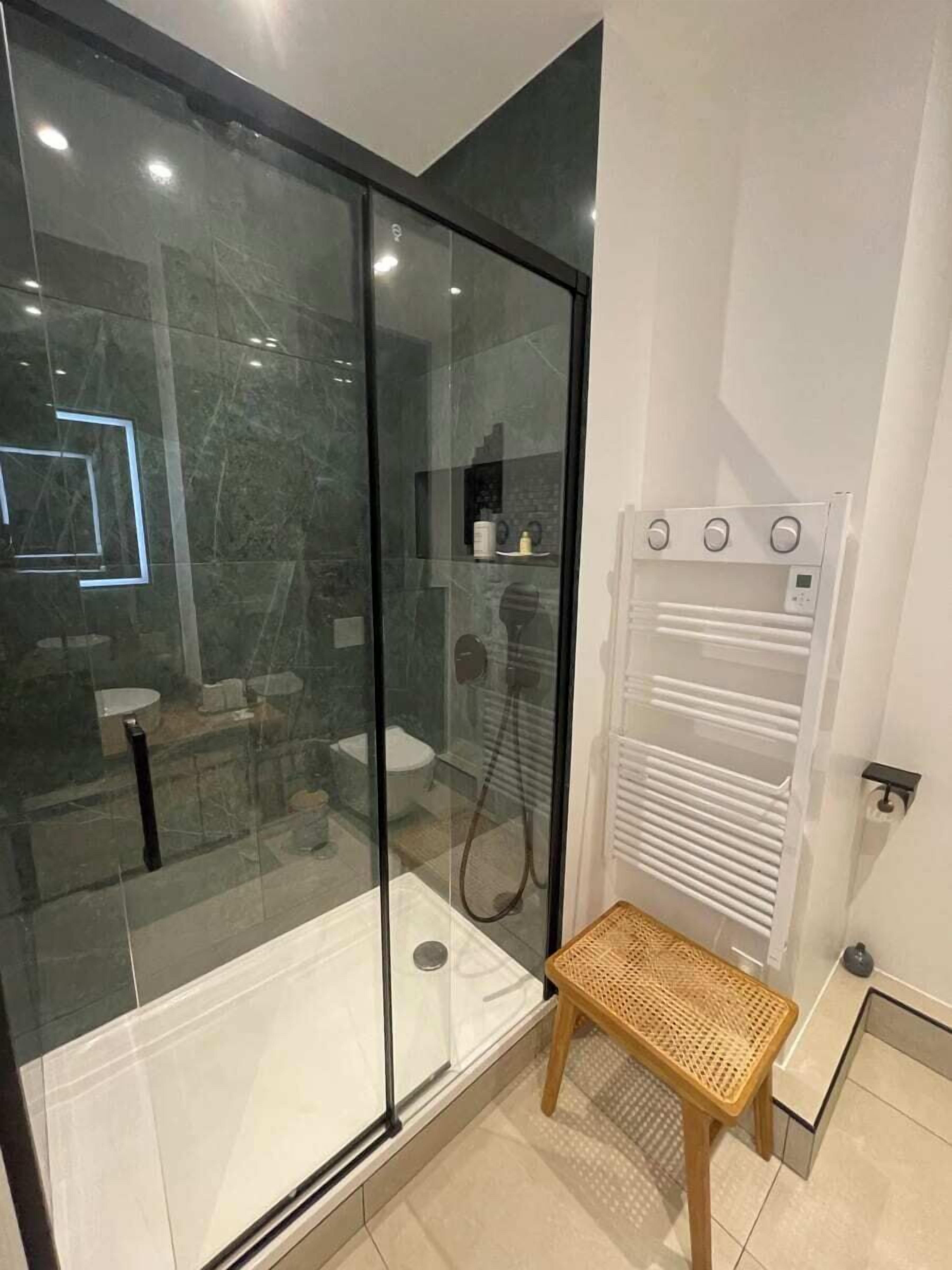 A modern bathroom with a glass shower enclosure, a white towel rack, and a small wooden stool.