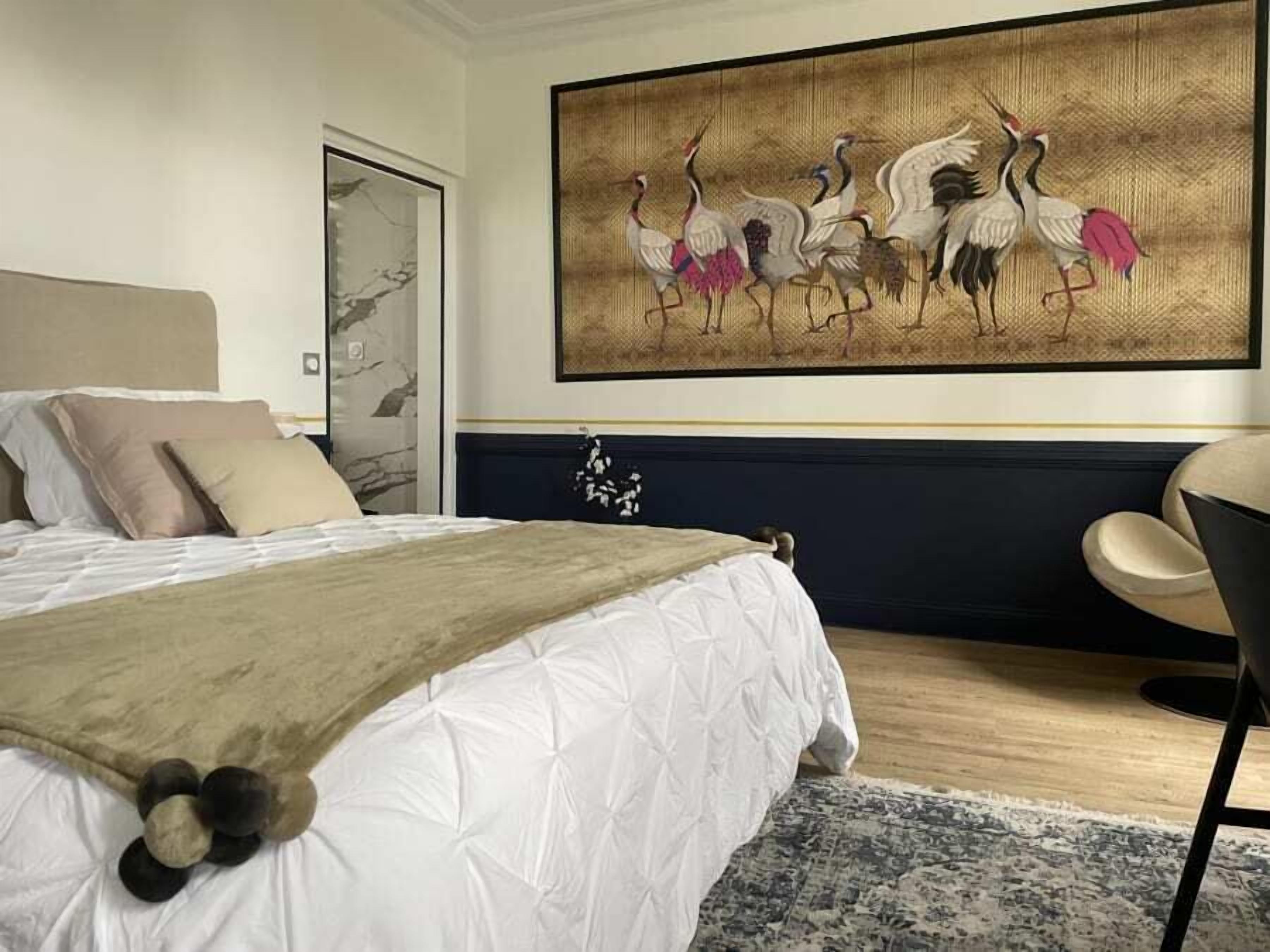A bedroom with a large bed, a headboard, a wall-mounted painting of birds, a chair, and a door.