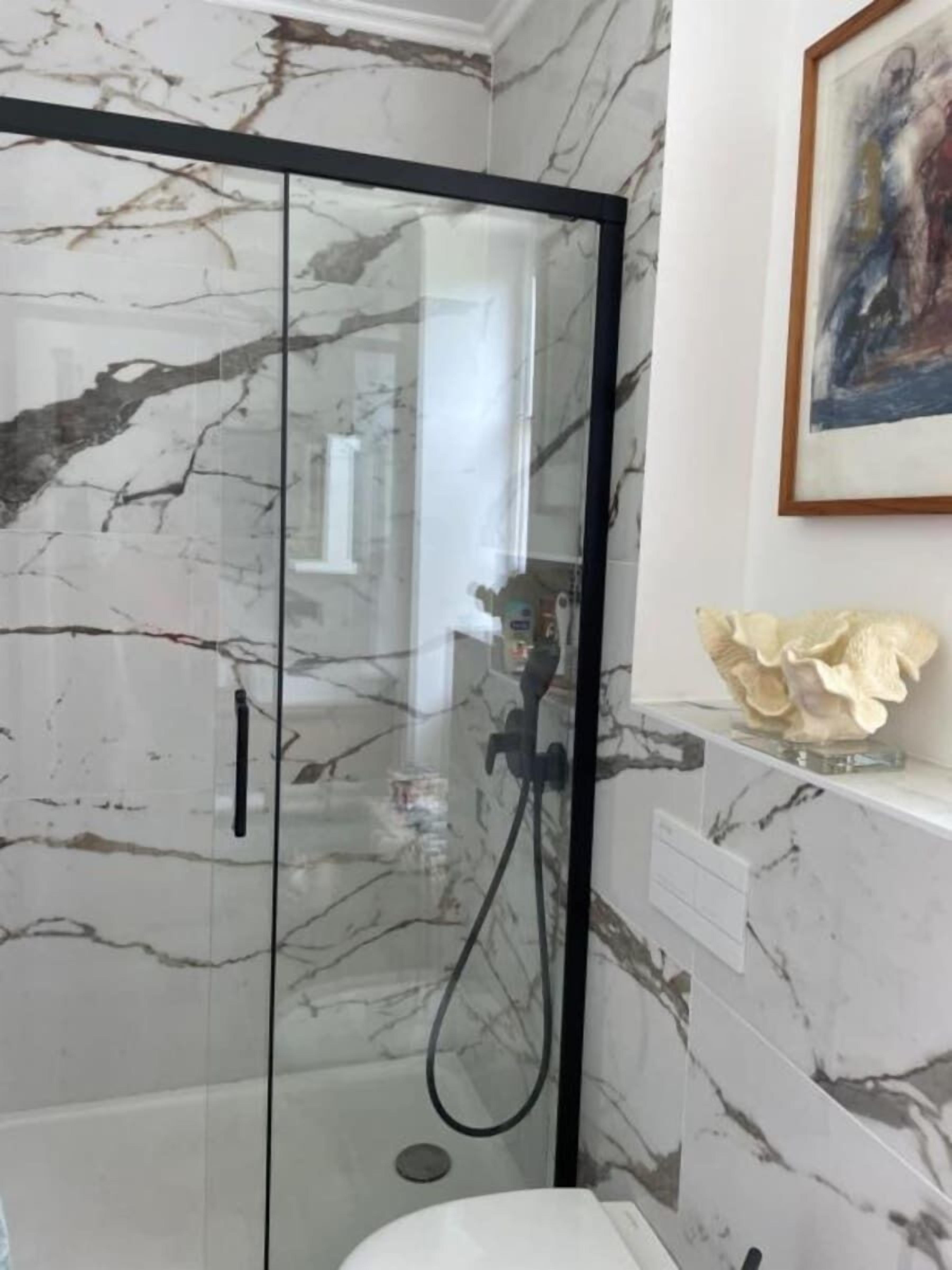 A bathroom with a glass-enclosed shower, marble walls, and a framed artwork above the sink.