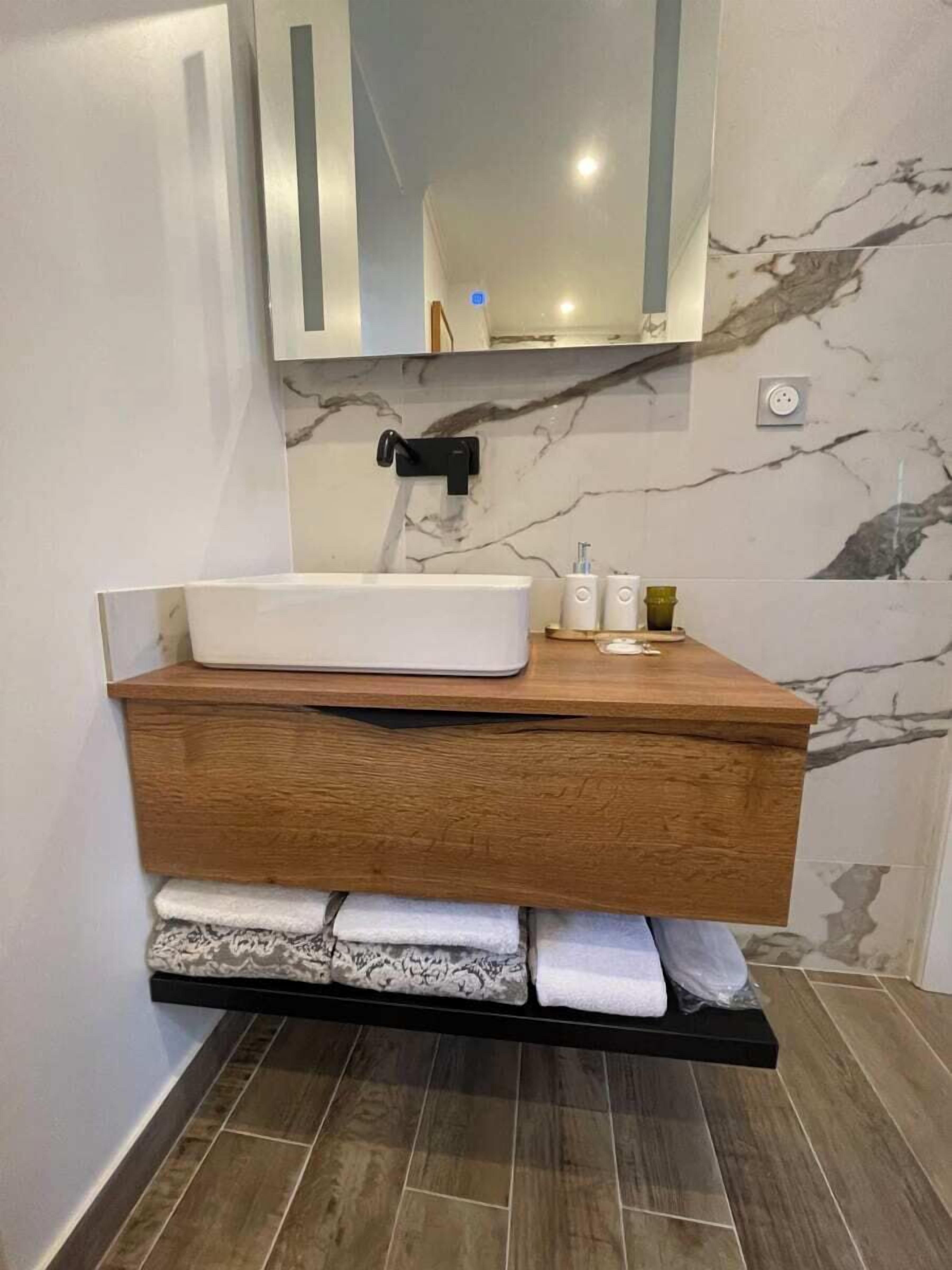 A modern bathroom with a marble backsplash, a wooden vanity, a built-in shelf with towels, and a mirror above the sink.