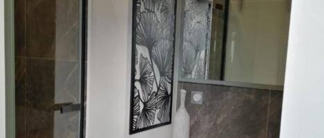 Un baño con cabina de ducha de vidrio, tocador de madera y un jarrón blanco, además de un motivo de hojas enmarcado en la pared.