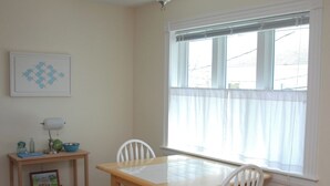 A dining area with a wooden table and white chairs, a small side table with a lamp, a framed picture, and a window with blinds. -  In-Town Bar Harbor, One Bedroom, Huckleberry Suit (Bar Harbor)