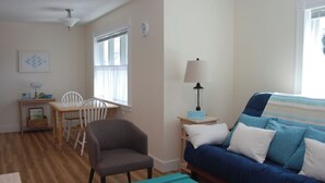 A cozy living room with a blue sofa, a wooden dining table with chairs, a lamp, and a window with curtains. -  In-Town Bar Harbor, One Bedroom, Huckleberry Suit (Bar Harbor)