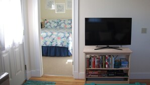 A room with a television on a wooden stand, a bookshelf with books and board games, a bed with a floral bedspread, and a door leading to another room. -  In-Town Bar Harbor, One Bedroom, Huckleberry Suit (Bar Harbor)